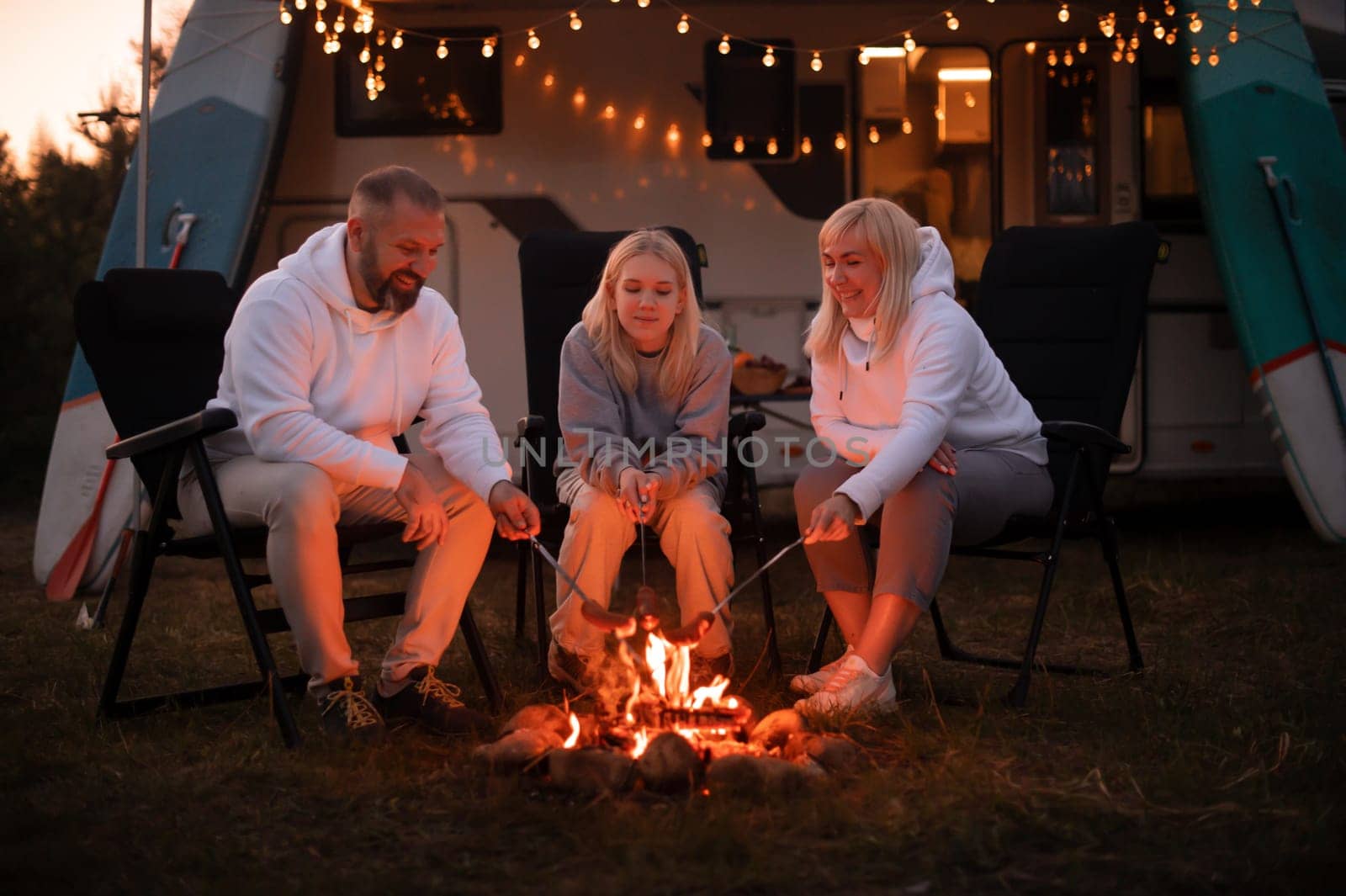 A family cooks sausages on a bonfire near their motorhome in the woods.