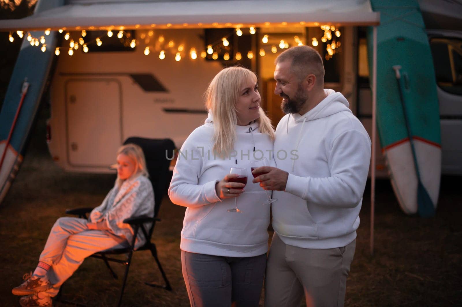 A married couple with glasses of wine stands against the background of a motorhome and rests together by the campfire. Evening family vacation.