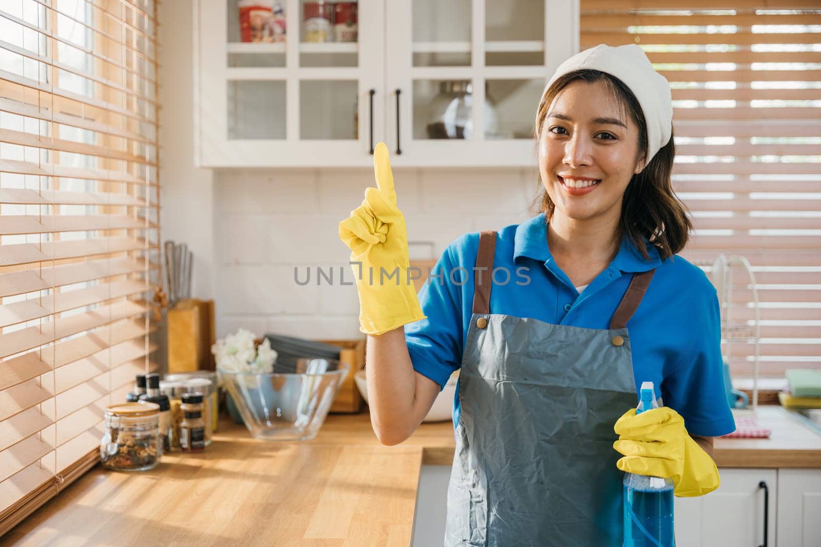 Housekeeping-ready woman in uniform with apron and glove holds spray bottle for home cleaning. Ensuring hygiene and cleanliness. Clean disinfect home care. maid with liquid. by Sorapop