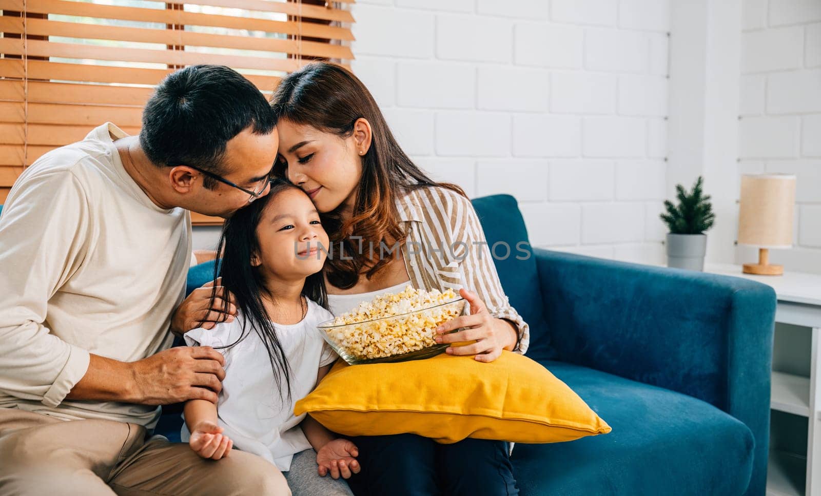 A happy family filled with joy watches TV while eating popcorn on modern sofa at home by Sorapop