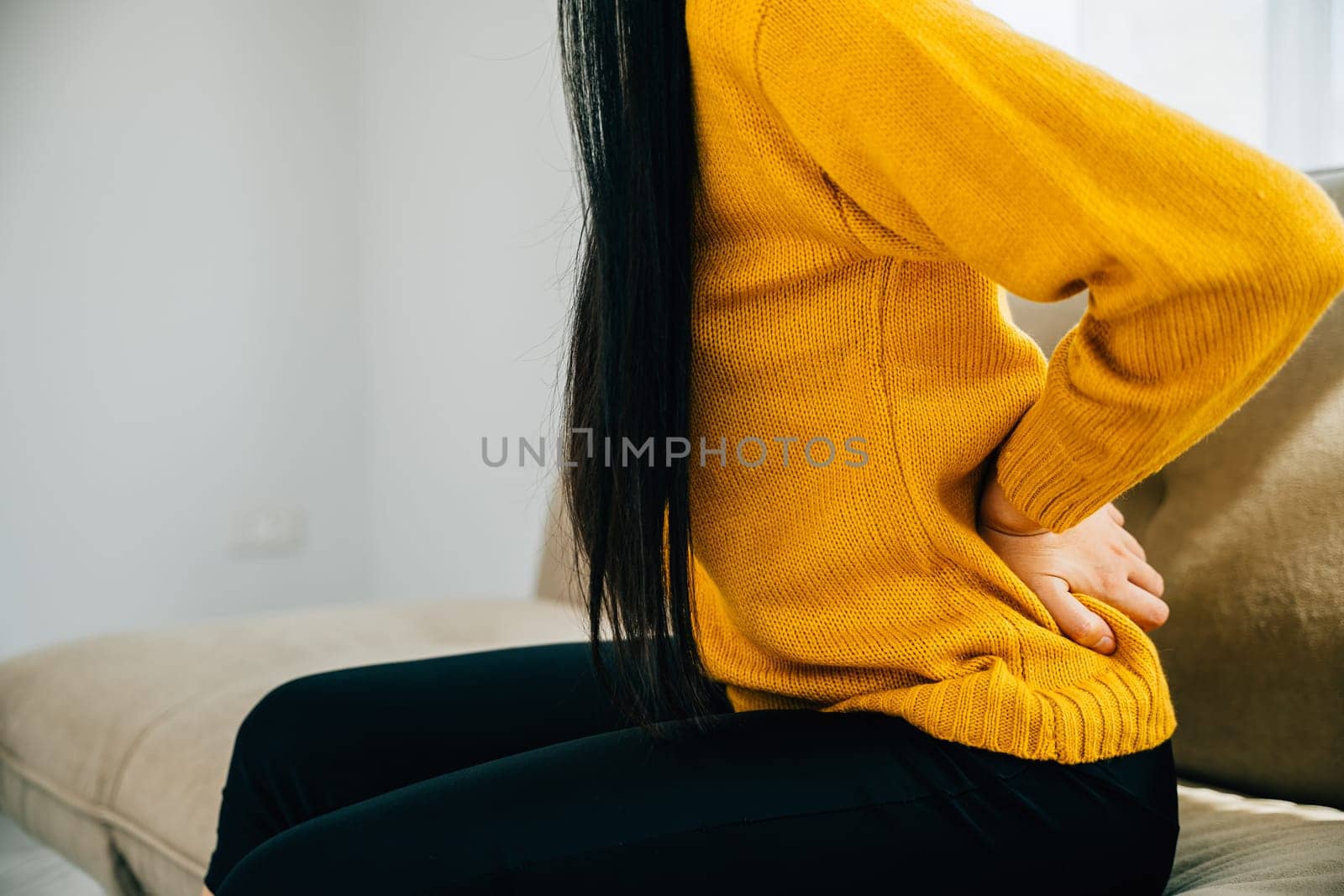 Highlighting unbearable back pain, Asian woman on sofa holds her lower back. Illustrating chronic backache discomfort and the need for medical care and attention. healthcare and problem concept