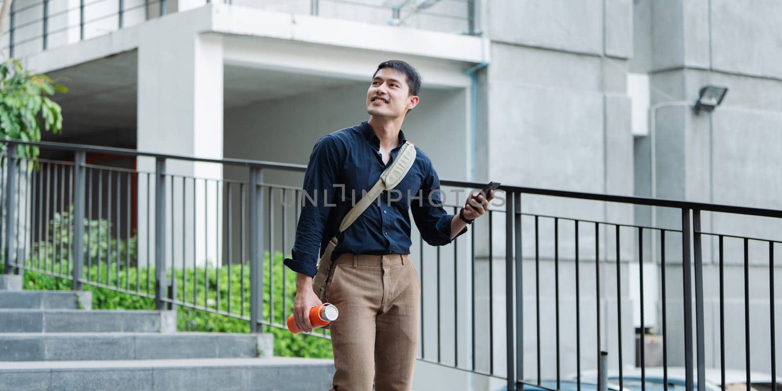 Smiling asian businessman hold reusable eco-friendly ecological cup and using mobile while sitting at park.