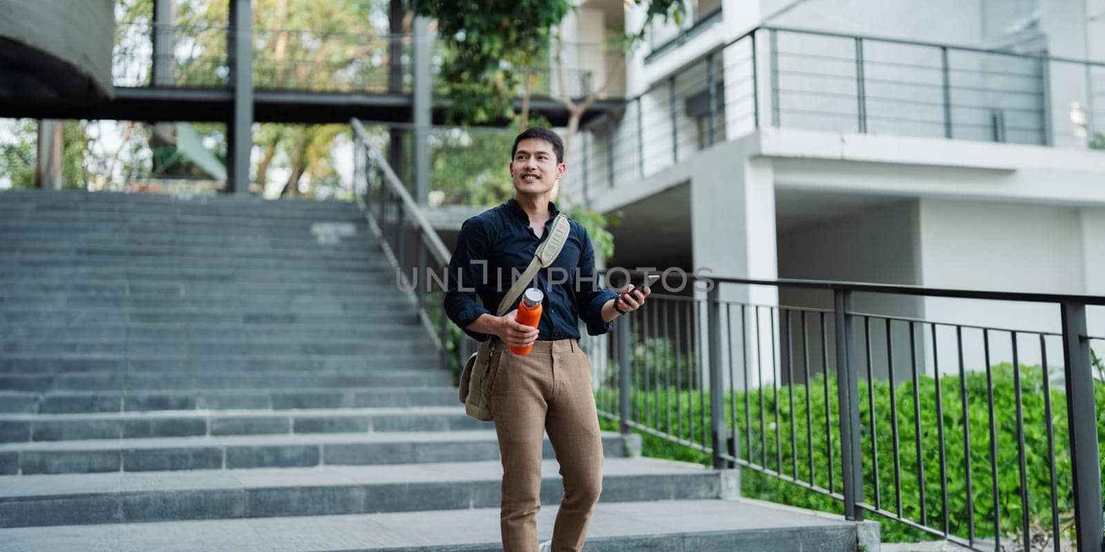 Smiling asian businessman hold reusable eco-friendly ecological cup and using mobile while sitting at park.