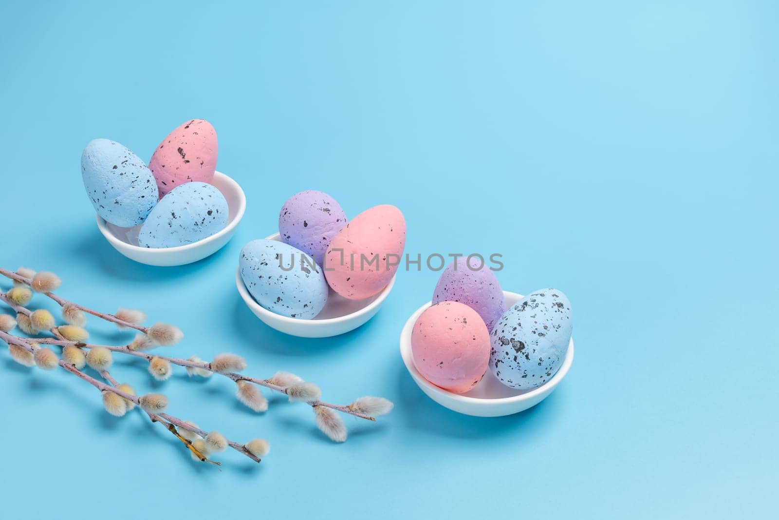Porcelain saucers with colored Easter eggs and willow branches with catkins on the blue background. Top view.