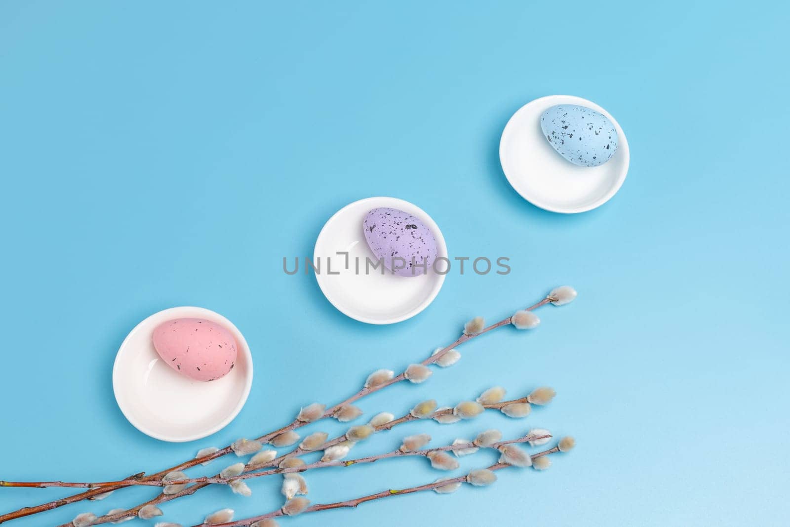 Porcelain saucers with colored Easter eggs and willow branches with catkins on the blue background. Top view.