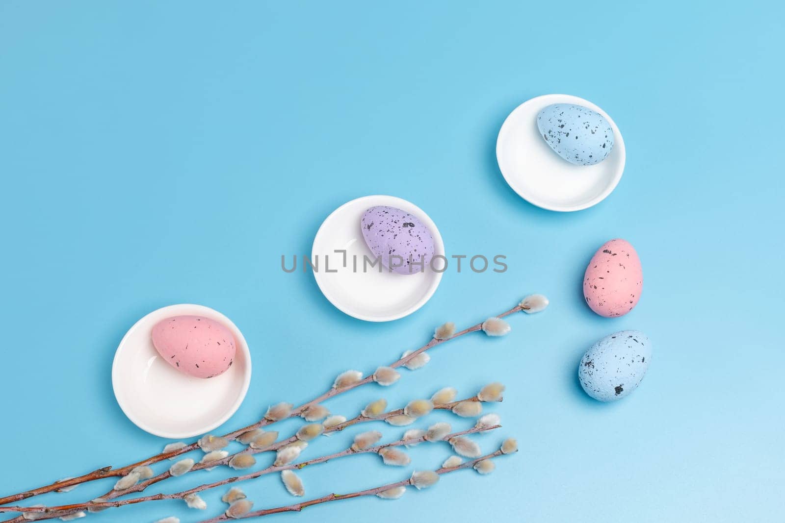 Porcelain saucers with colored Easter eggs and willow branches with catkins on the blue background. Top view.