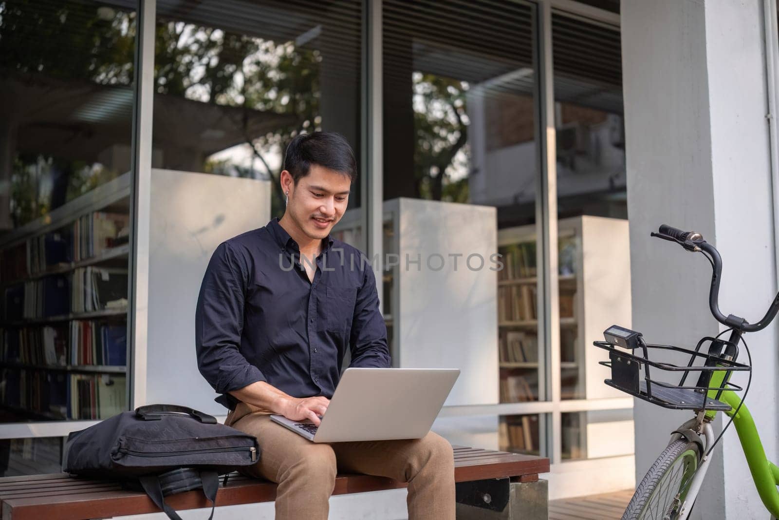 A young Asian businessman rides a bicycle to work. Sitting outside the office working using a laptop. by wichayada