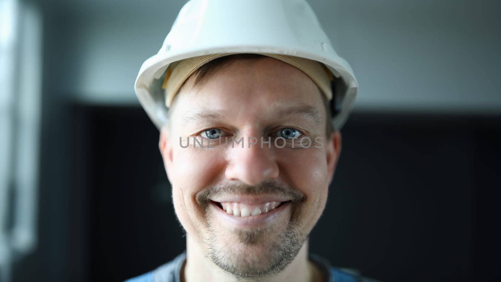 Close-up of cheerful worker face. Macro shot of foreman in protective white helmet posing on camera indoors. Happy handyman. Renovation and interior designer concept