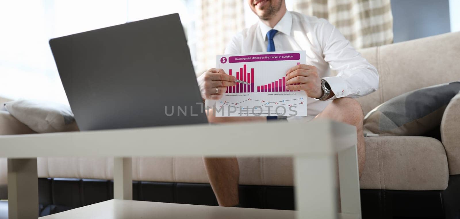 Close-up of male hands holding financial statistic and talking on telework application. Male in pants on sofa indoors. Remote conference on computer. Business and work from home concept