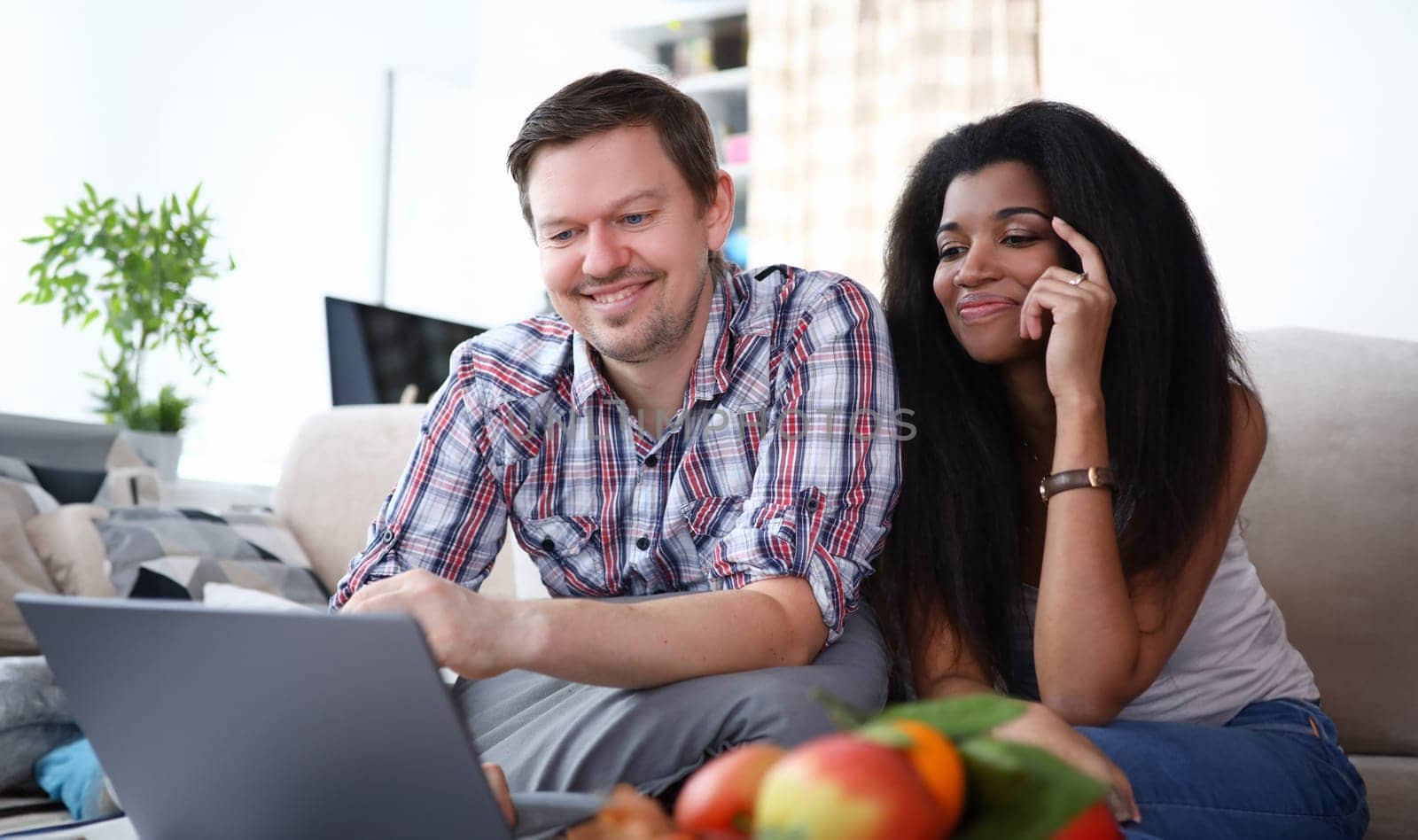 Portrait of smiling cute couple searching for information on laptop. Man and afro-american woman sitting on sofa in living room. Leisure together. Technology and love concept