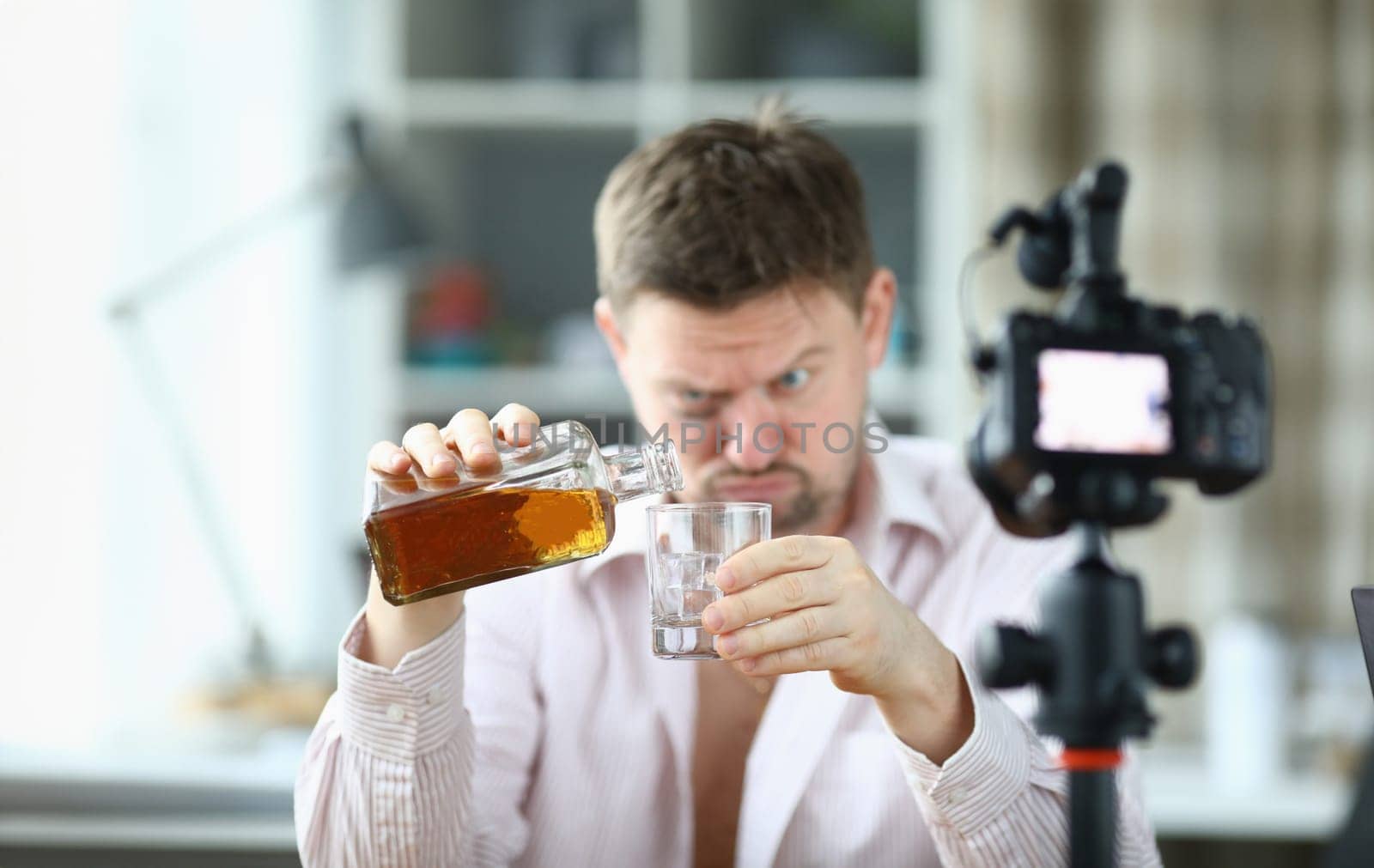 Portrait of man pouring whiskey into glass from bottle. Guy looking drunk and filming on videocamera. Unbutton shirt and bruise under eye. Production on camera