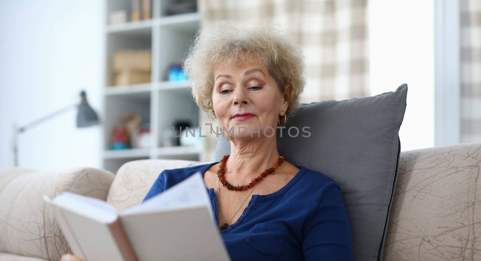 Portrait of concentrated smiling female reading hardcovered book. Good-looking attractive senior woman resting on couch at home. Spare time and hobby concept