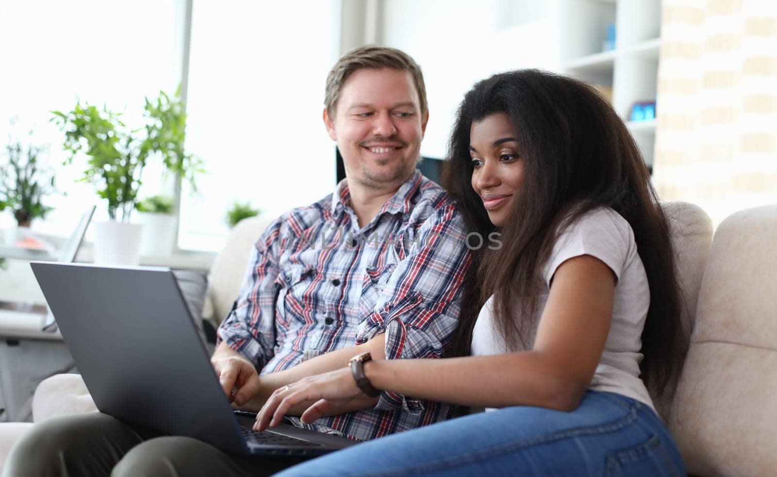 Portrait of couple sitting on sofa and choosing what film to watch. Afro-american young woman using laptop. Happy smiling middle-aged man. Relationship and quality time concept