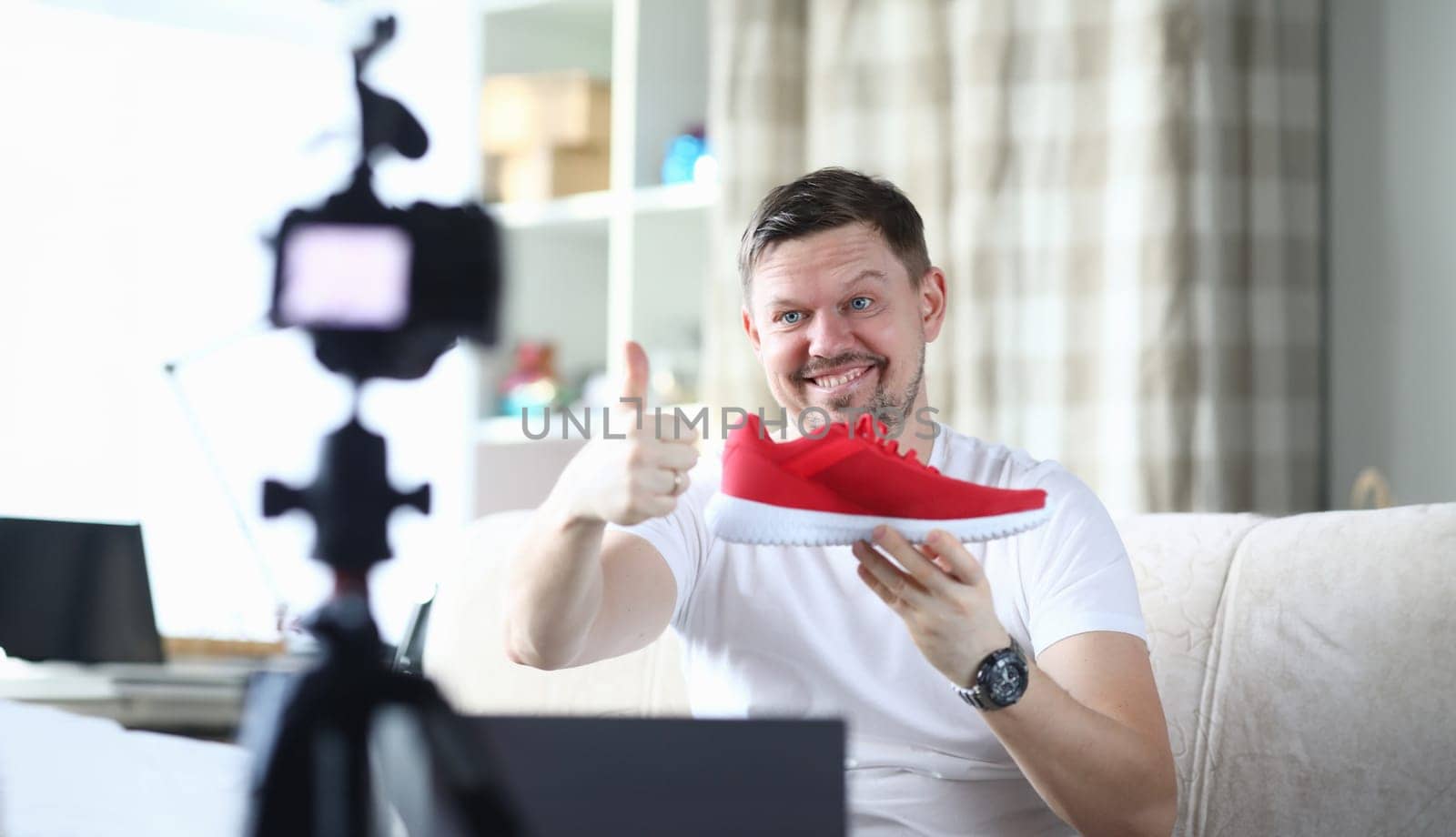 Portrait of smiling middle-aged man filming red sneaker on camera and tell in vlog. Male describing quality of new boots on display of digital device. Technology concept