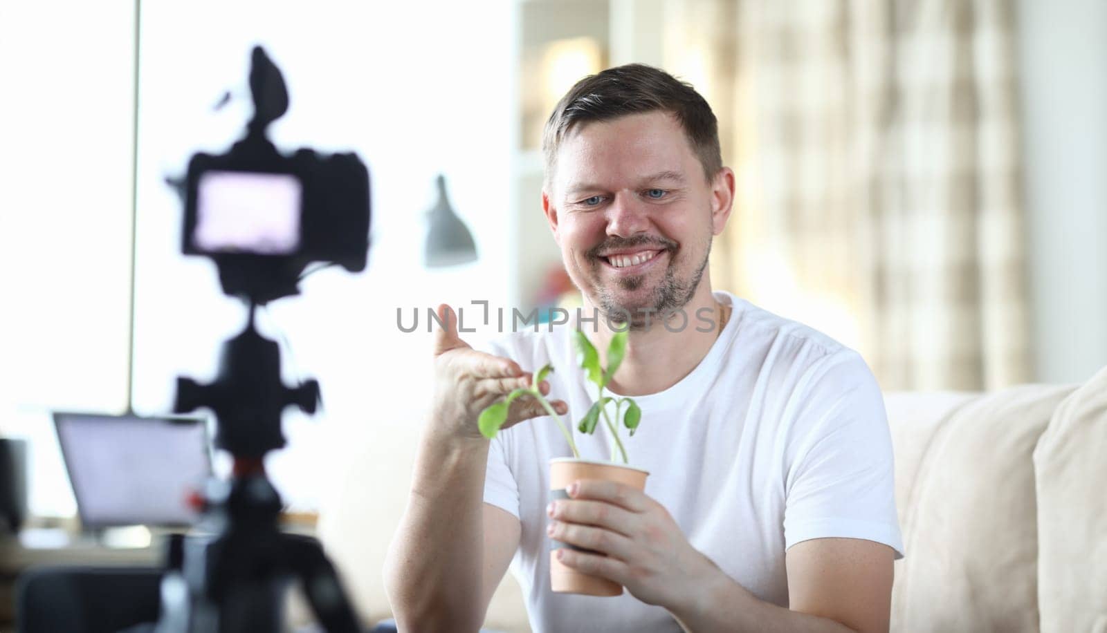Portrait of middle-aged male smiling and showing on video camera plant in pot. Man beginning to film for vlogs. Beginner blogger and modern technology concept