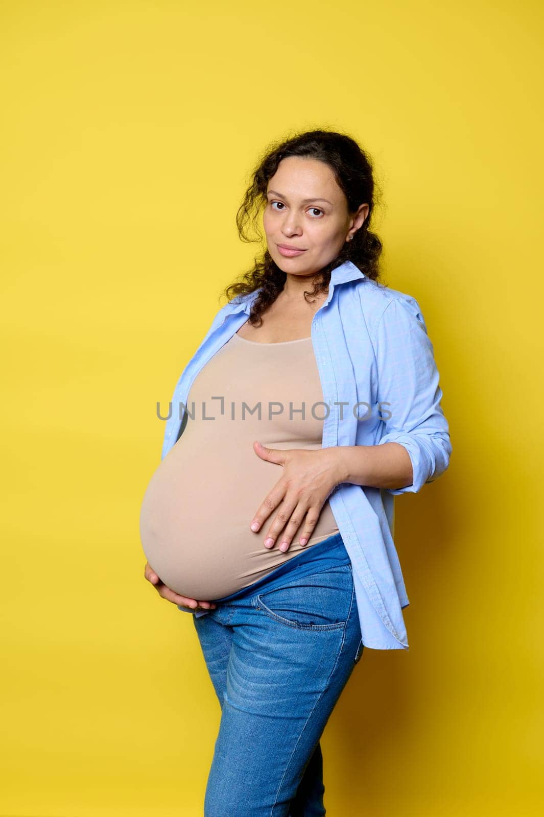 Confident young pregnant woman touches her belly in last trimester of happy pregnancy, isolated yellow studio background by artgf