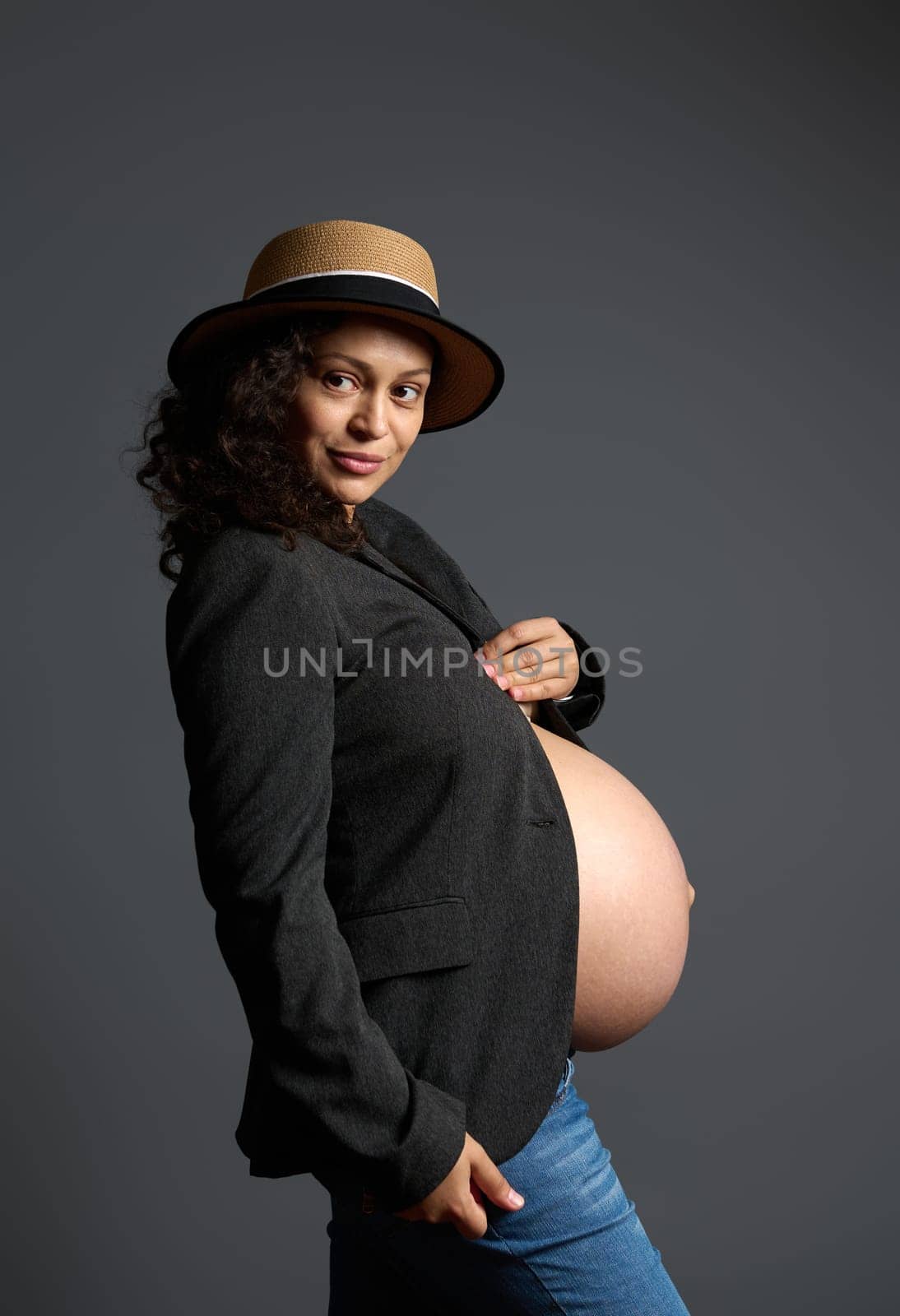 Attractive and pensive young pregnant woman, expecting baby, holding her beautiful big belly, dreamily looking aside a copy ad space on fashion gray backdrop, dressed in gray blazer and blue jeans