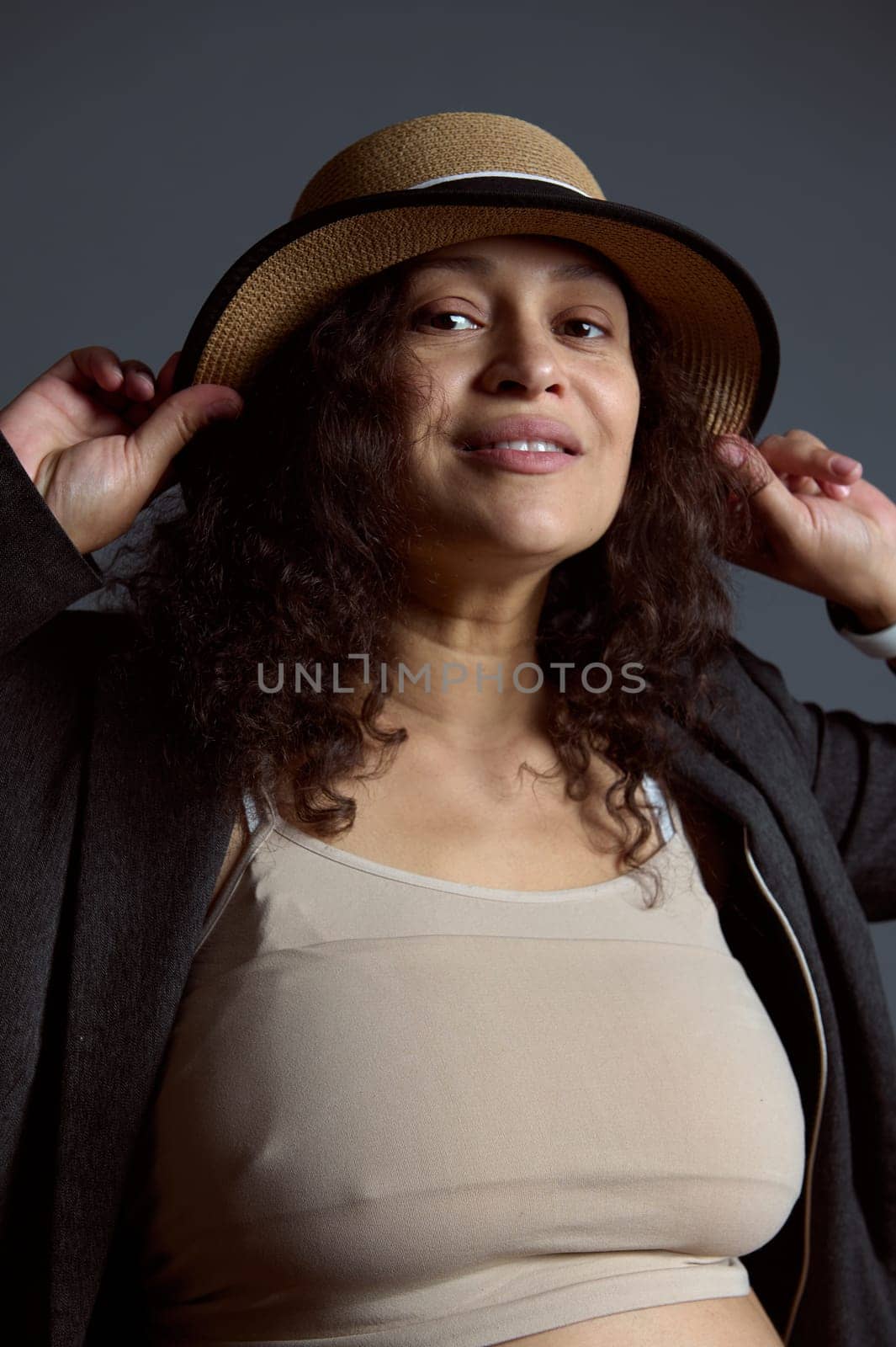 Authentic pregnant woman in beige top, gray blazer and a straw hat, smiling at camera, over fashion gray background by artgf