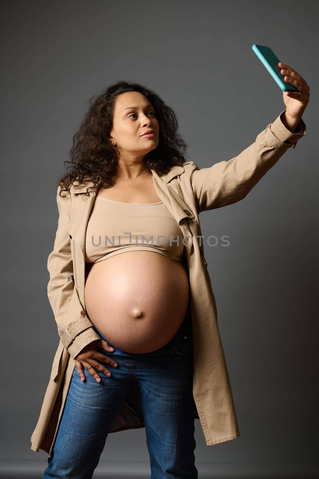 Stylish curly pregnant woman dressed in blue jeans and trendy trench, taking a selfie on mobile phone, sharing her lifestyle in last trimester of pregnancy on social media, isolated over gray backdrop