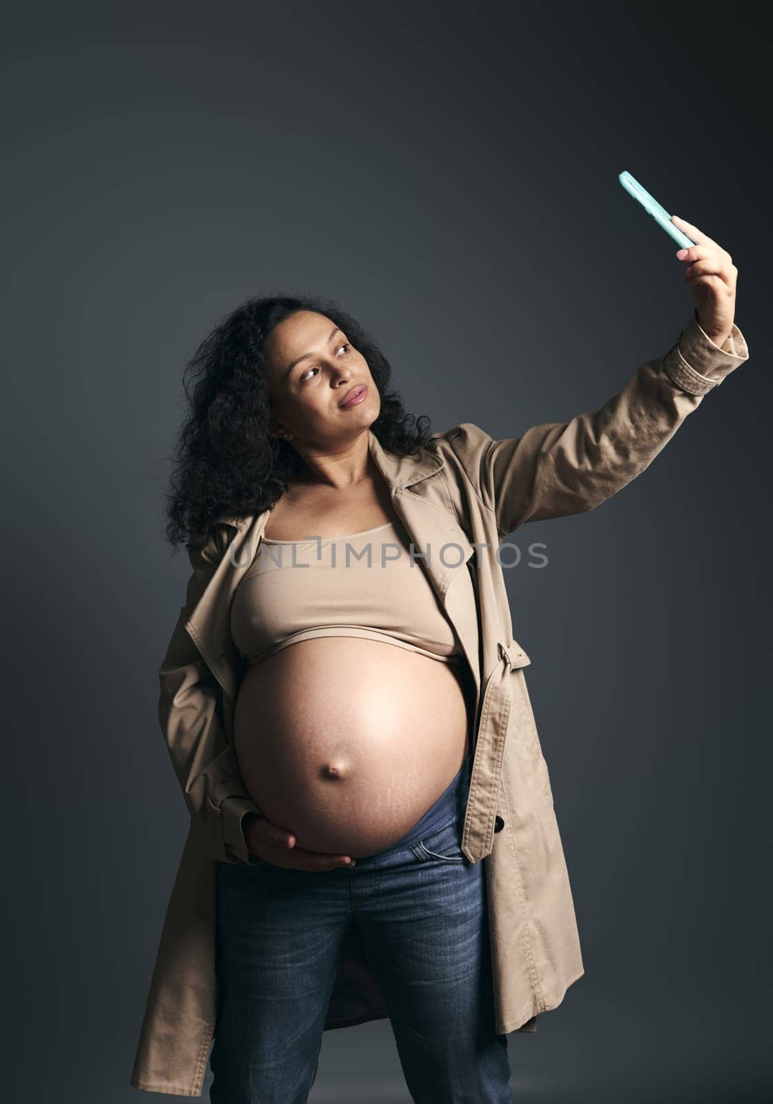 Happy pregnant woman takes self portrait on mobile phone while strokes her big belly, shares her lifestyle in last trimester of pregnancy on social media, isolated over fashion gray studio background