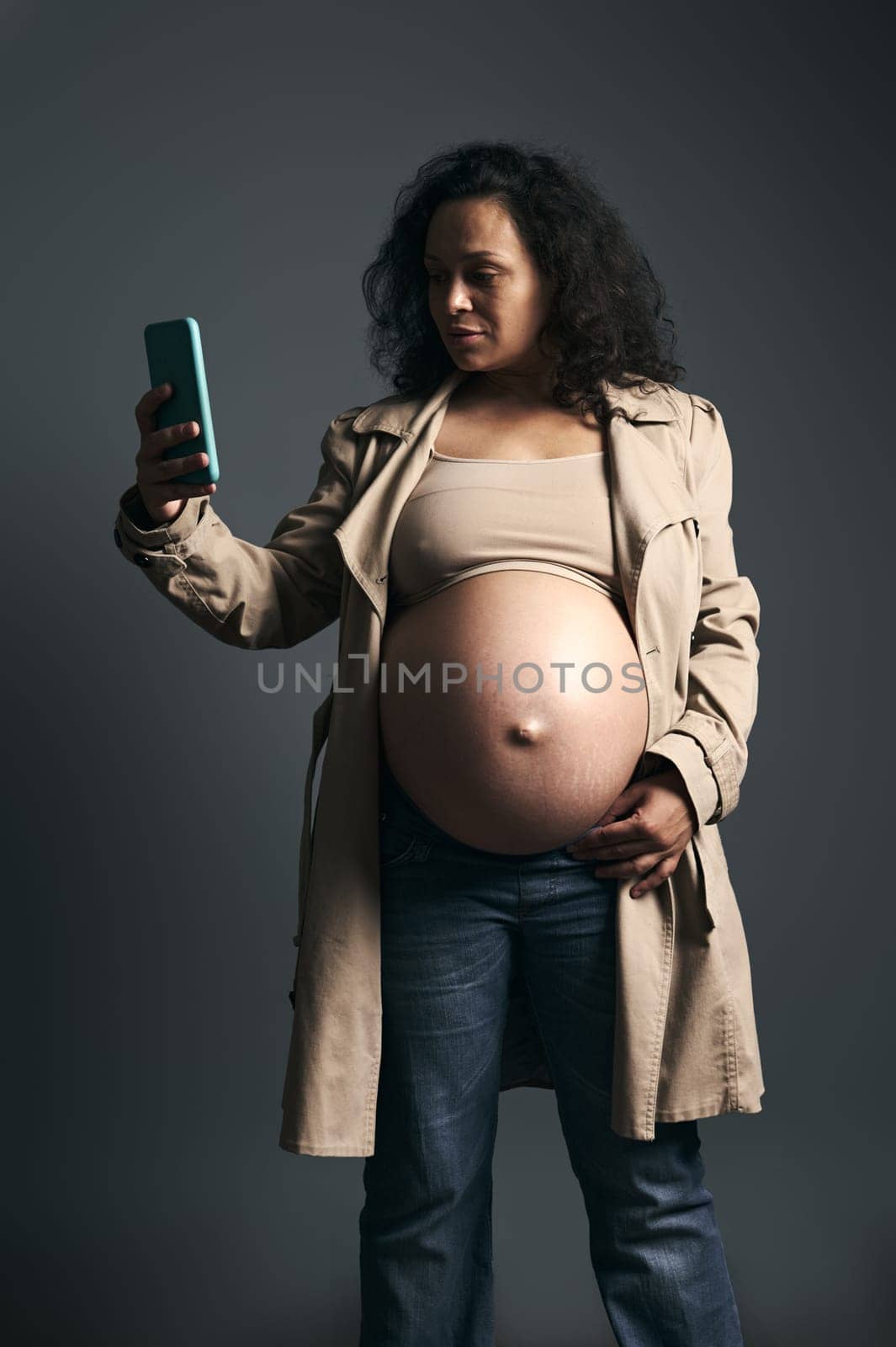 Pregnant woman with naked belly, in stylish beige trench, using smart mobile phone, isolated over gray studio background by artgf