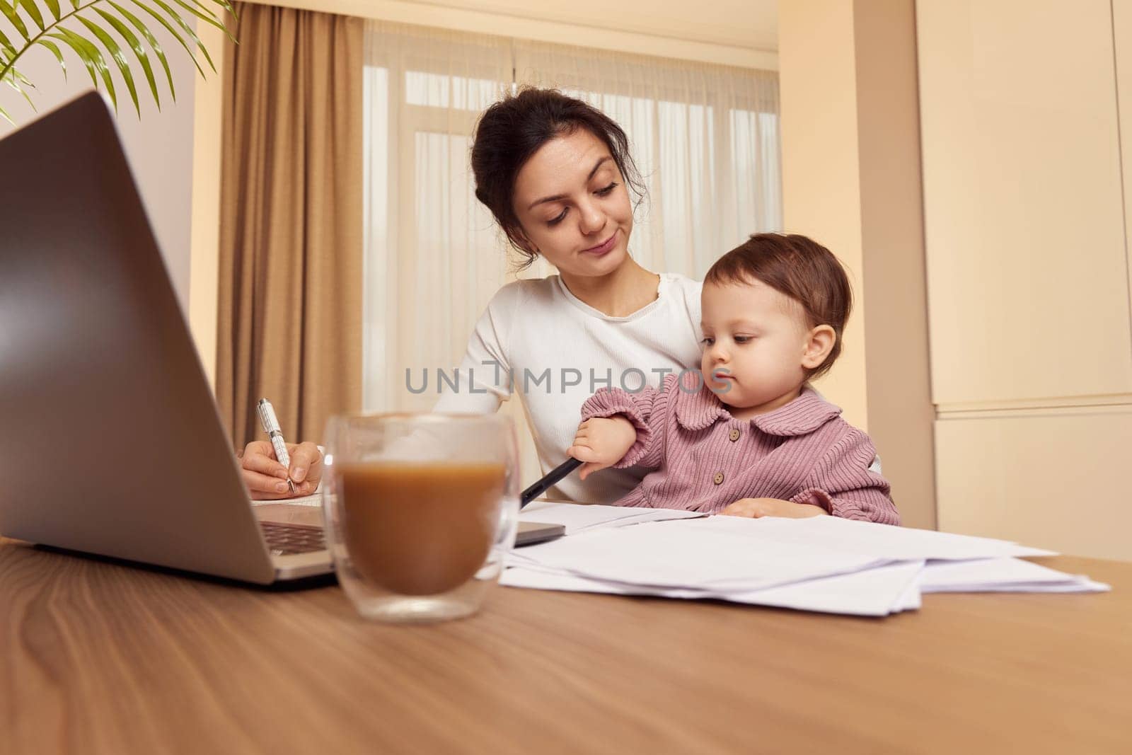 tired woman working on laptop at home with her little baby girl. Child makes noise and disturb mother at work.