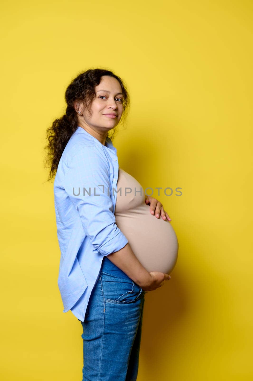Side portrait of a happy pregnant woman putting hands on her big belly in last trimester of pregnancy, smiling looking at camera, isolated over yellow studio background. Maternity leave concept