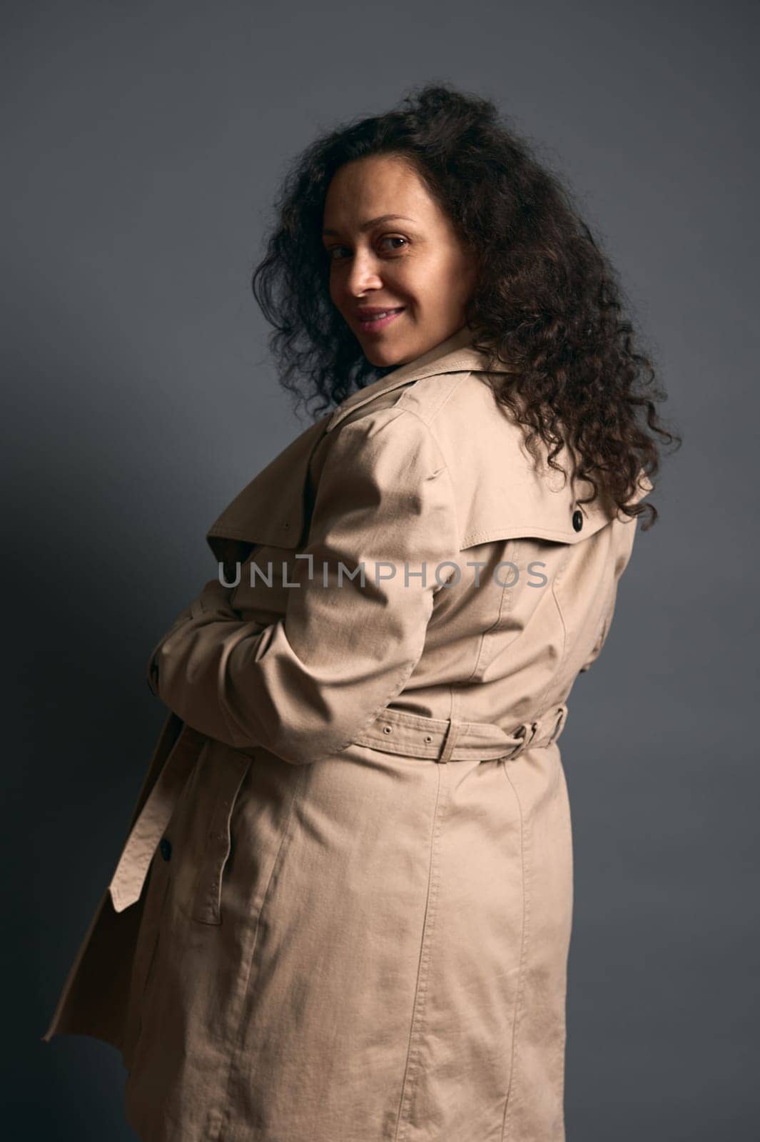 Authentic portrait of beautiful curly haired gravid woman in late time of pregnancy, wearing beige trench, smiling looking at camera through her shoulder, isolated over fashion gray studio background