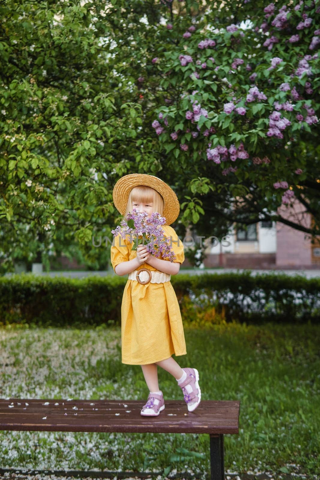 A little girl in a yellow dress and straw hat wearing a bouquet of lilacs. A walk in a spring park, blossoming lilacs