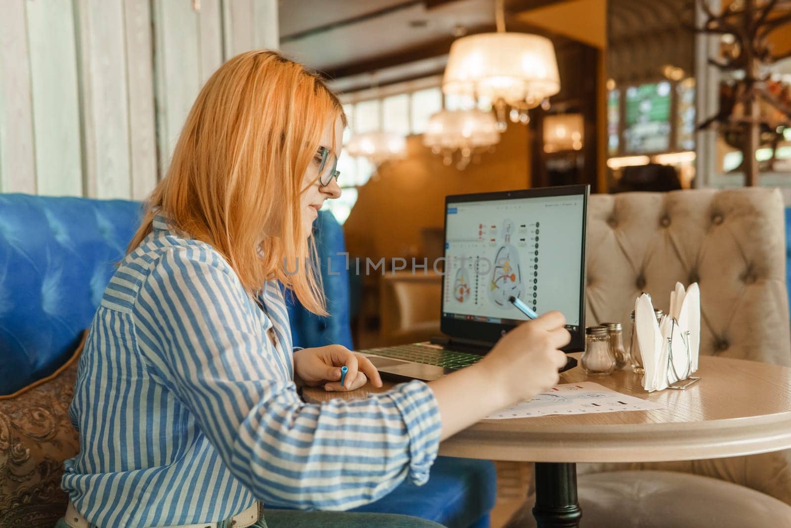 Tver, Russia-august 2, 2021. A woman in a cafe at a table with a laptop is studying the design of a person. The concept of studying esoteric sciences. A red-haired woman with glasses studies a rave card by human design.
