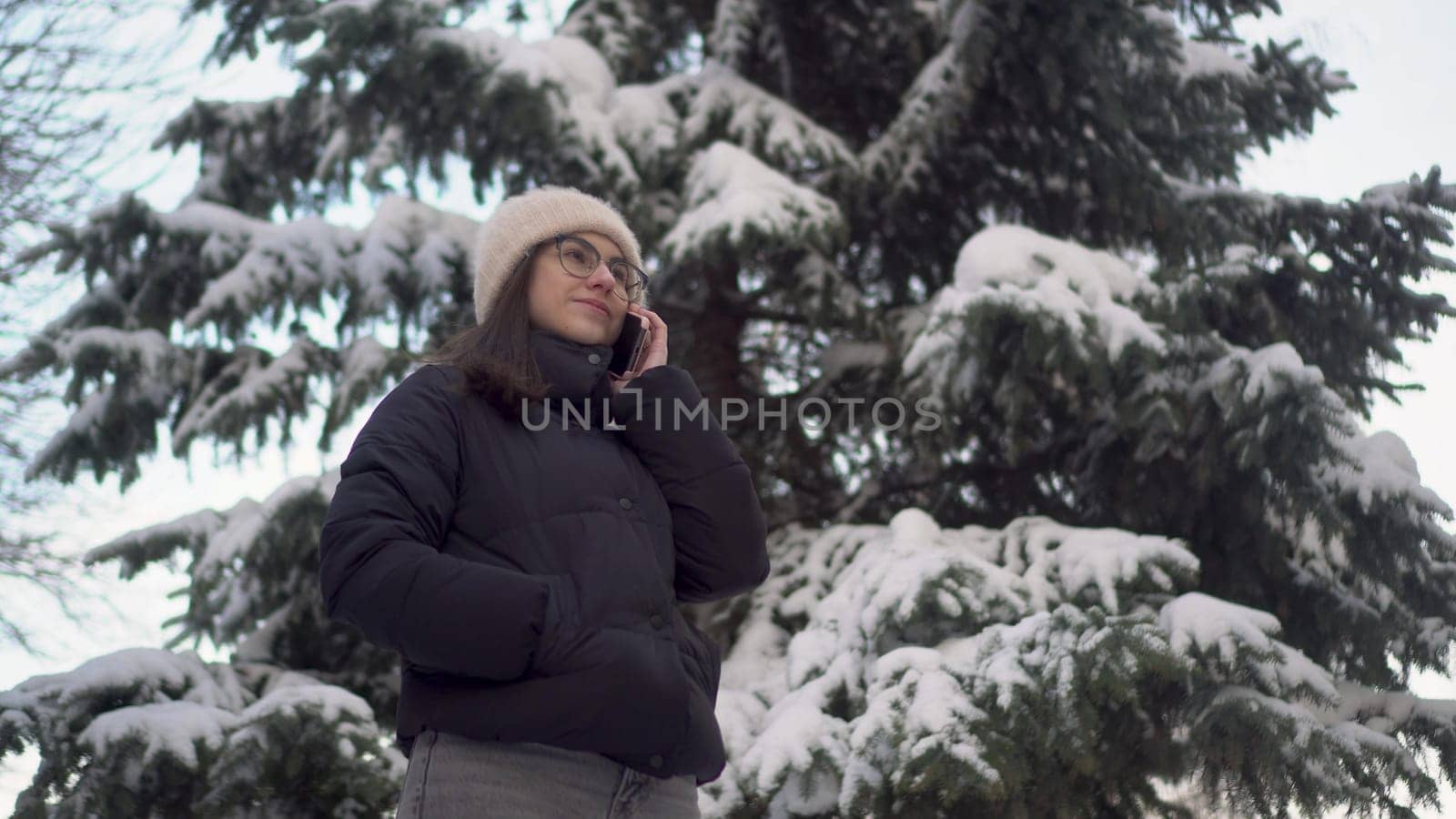 A young woman speaks on the phone near a tall spruce tree in winter. A girl with glasses communicates via cell phone against the backdrop of snowy spruce branches. 4k