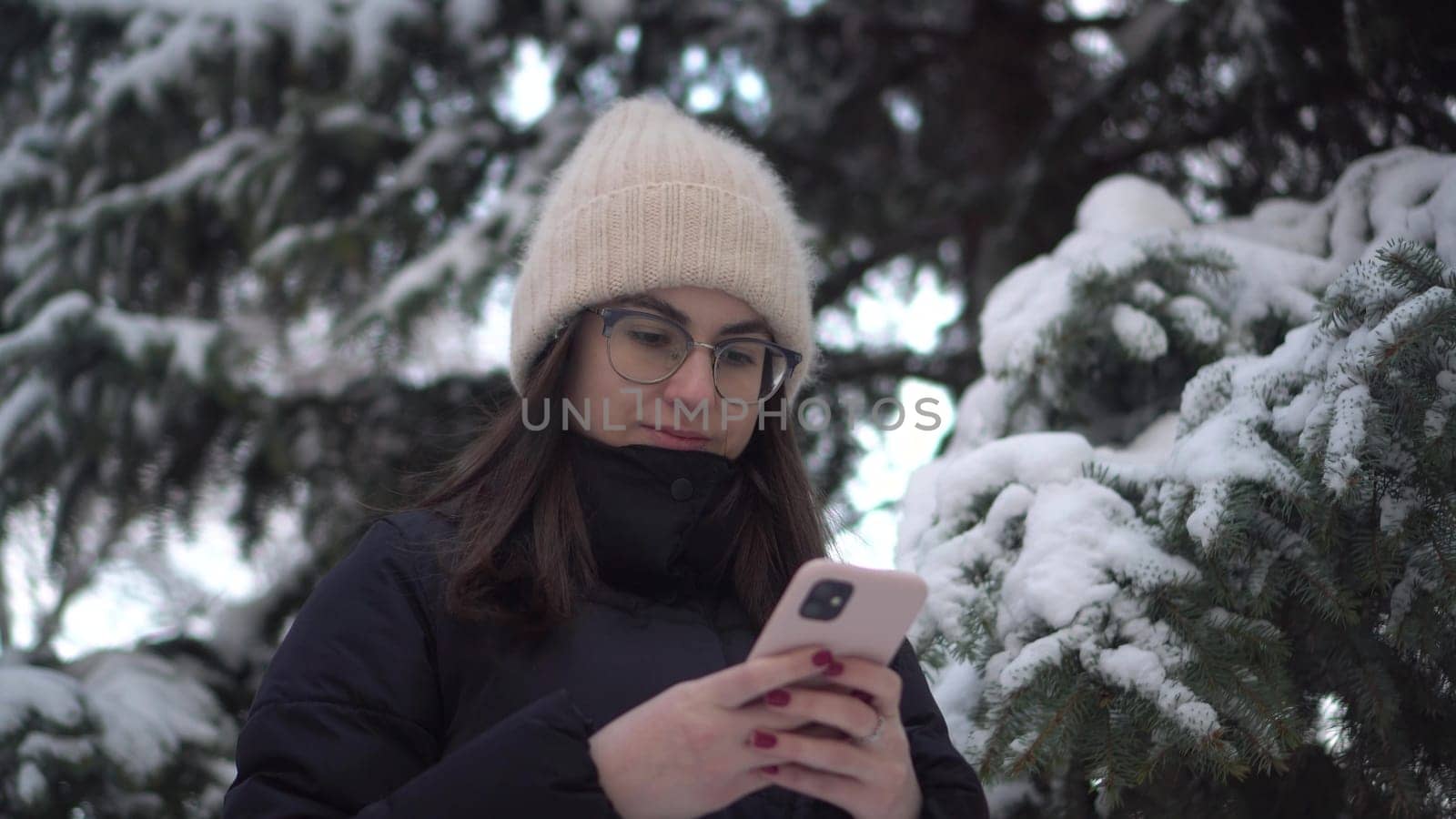 A young woman stands with a phone against a background of spruce in winter. A girl in glasses with a smartphone chats against the backdrop of snowy spruce branches. 4k