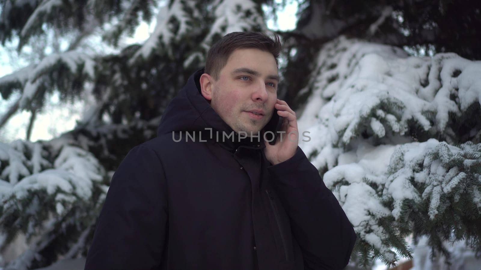 A young man speaks on the phone while standing against a background of spruce in winter. A man with a phone against a background of snowy spruce branches. 4k