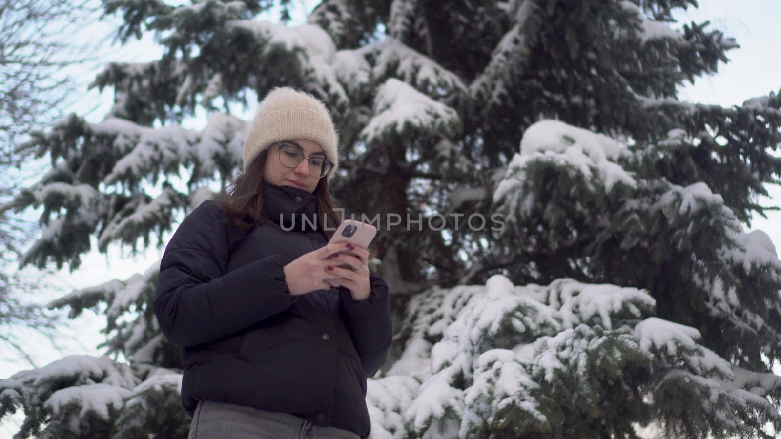 A young woman stands with a phone against a background of spruce in winter. A girl in glasses with a smartphone chats against the backdrop of snowy spruce branches. 4k
