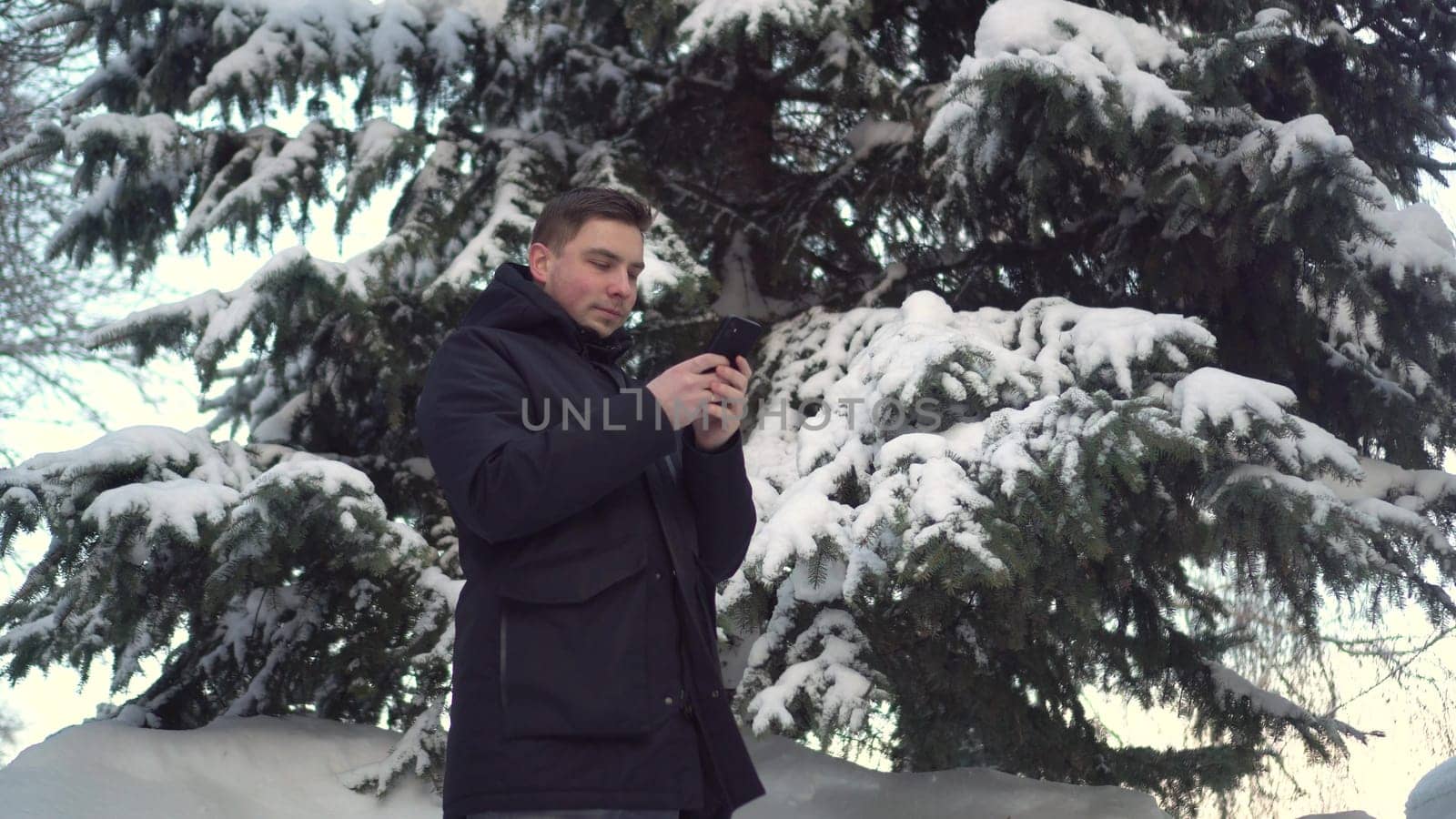 A young man stands on the phone against a background of spruce in winter. A man with a smartphone chats against the backdrop of snowy spruce branches. by Puzankov