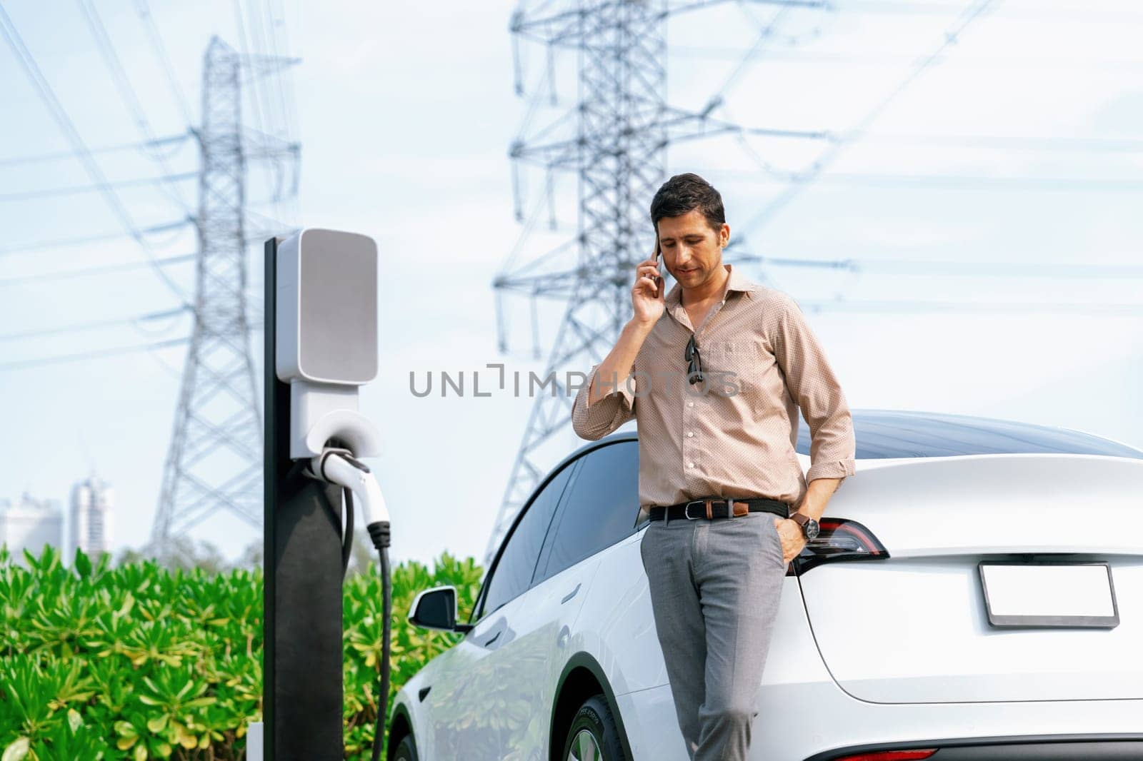 Man talking on the phone while recharge EV car battery at charging station connected to power grid tower electrical as electrical industry for eco friendly car utilization.Expedient