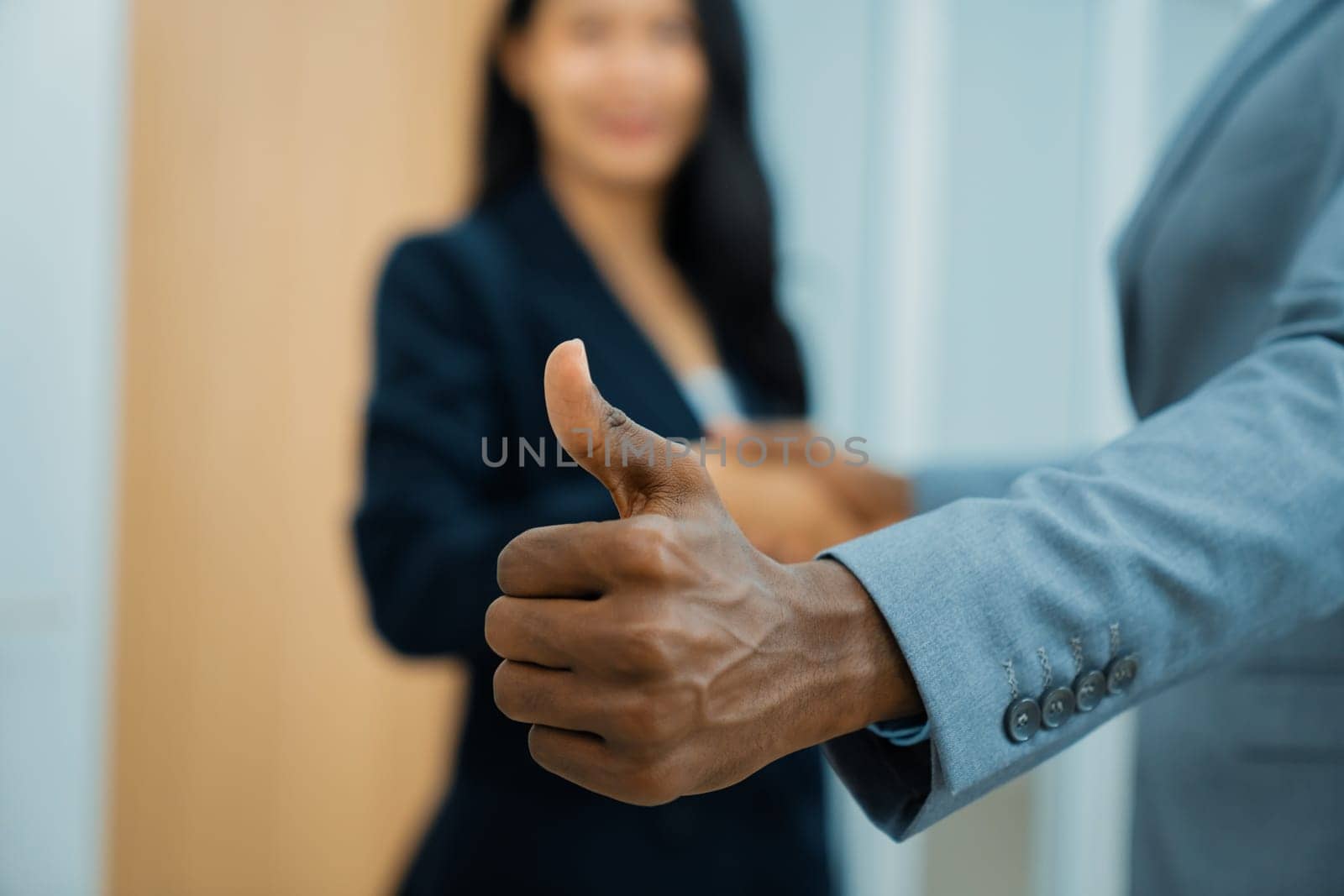 Closeup image of african businessman hand showing thump up to camera while young businesswoman standing behind. Represent recommend, support, good, positive, professional, reliable. Ornamented.