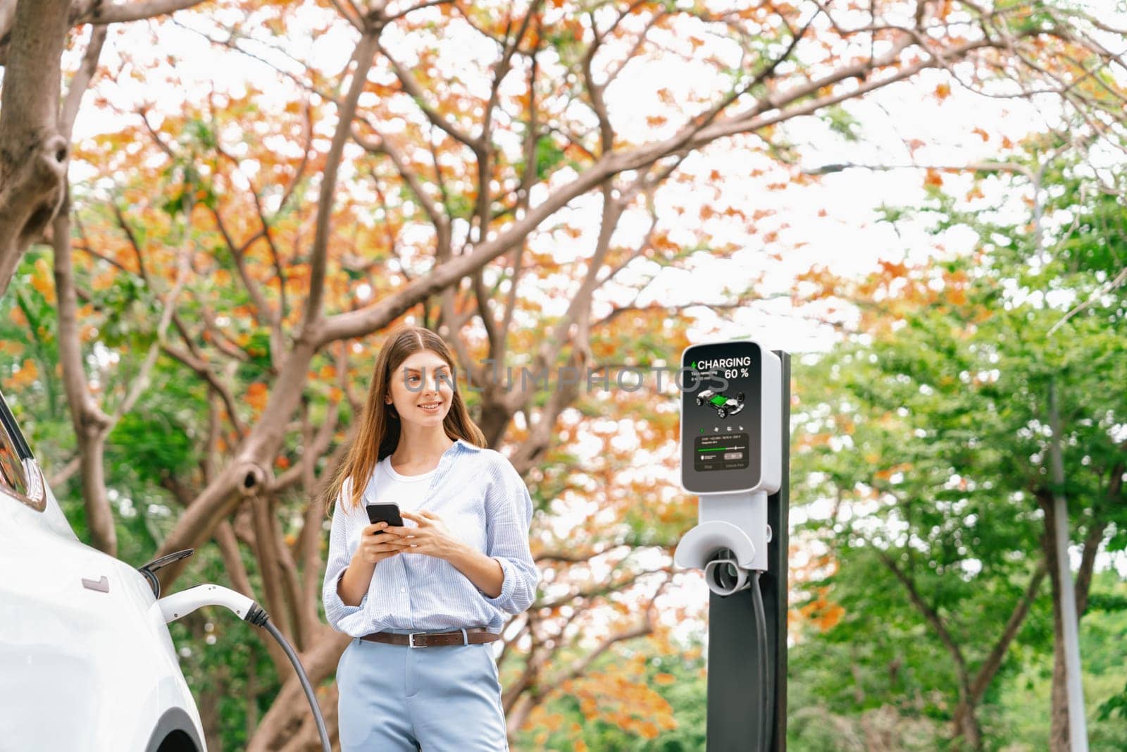 Young woman using smartphone online banking application to pay for electric car battery charging from EV charging station during autumn vacation holiday trip at national park or autumnal forest. Exalt