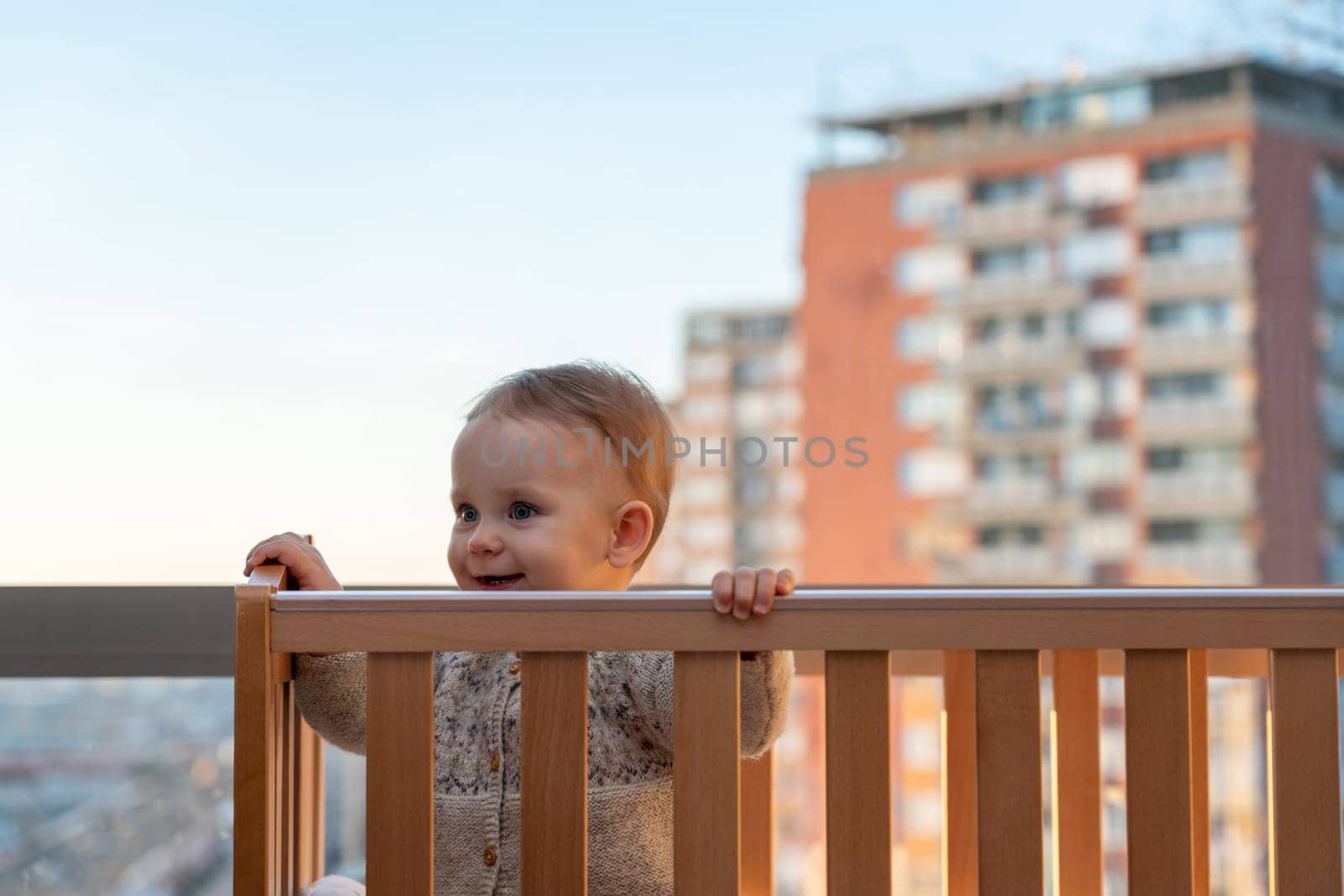 baby smiling cheerfully looks out of the crib against the backdrop of the city landscape.