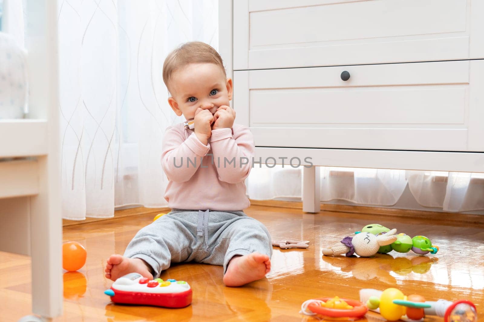 The baby hides his smile with his hands while sitting on the floor and playing with toys.