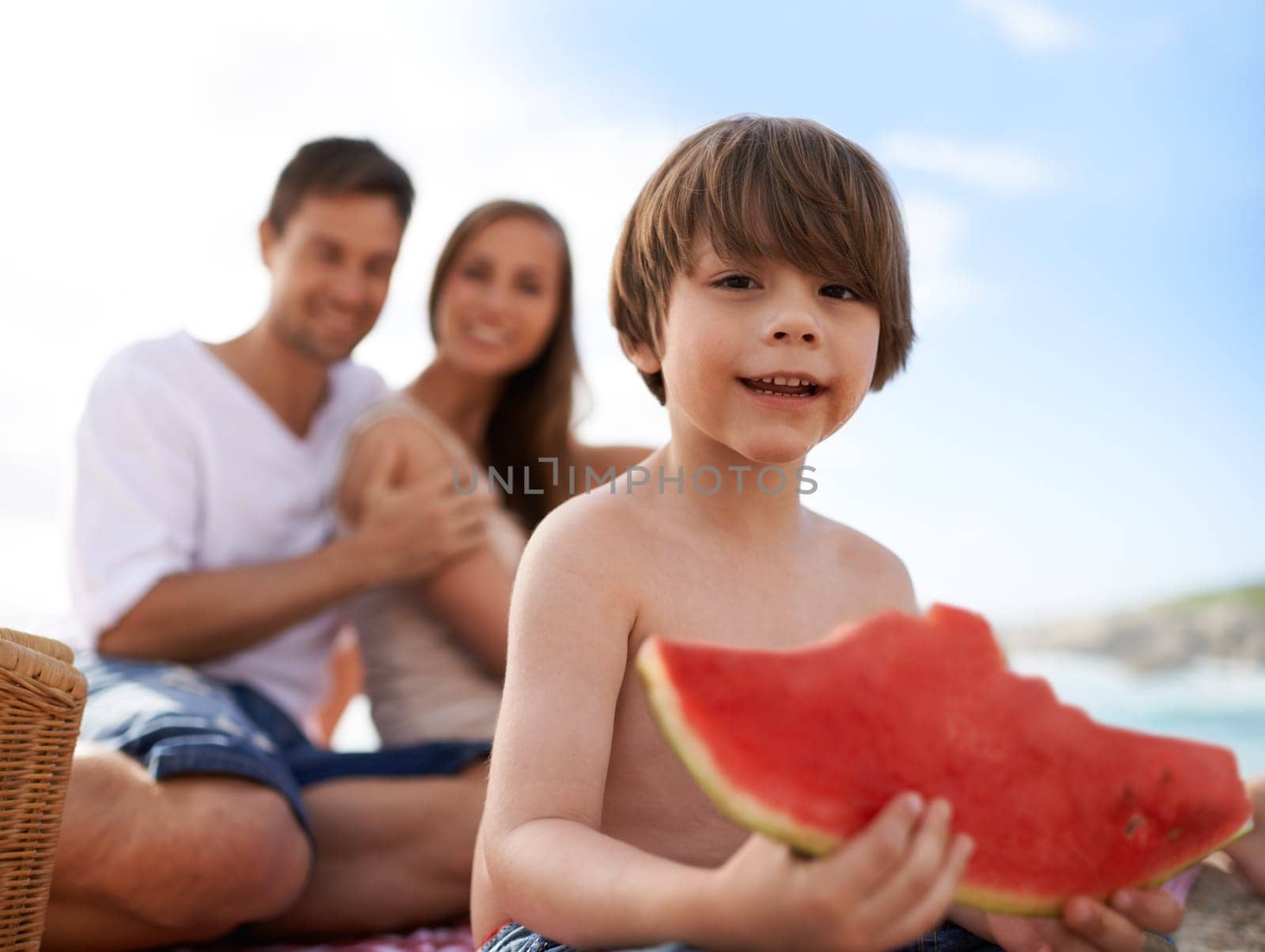Portrait, parents and kid on beach with watermelon, picnic and tropical holiday in Australia. Mother, father and happy face of boy child relax at ocean with sunshine, eating fruit and smile in summer.