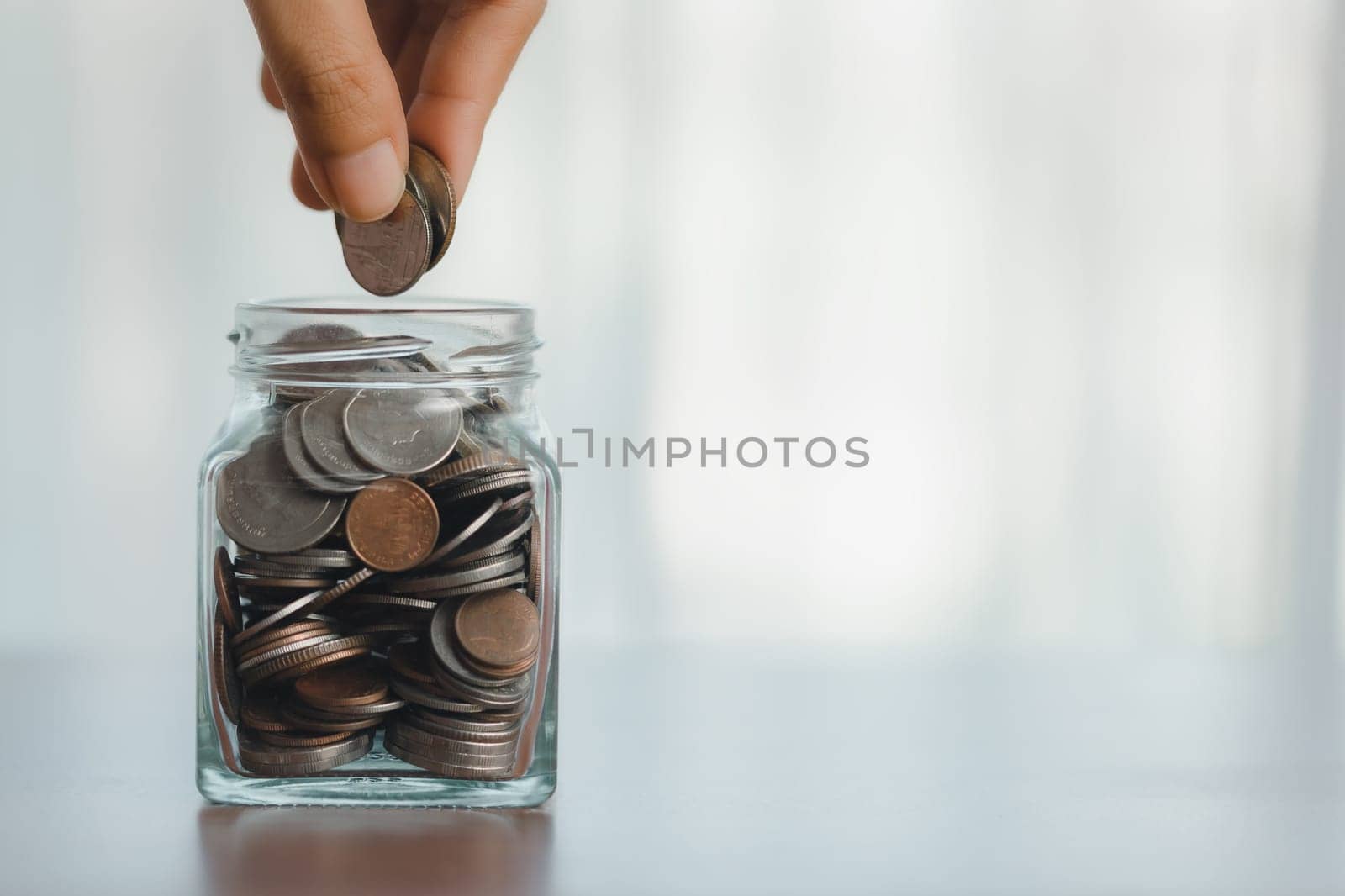 Glass jar with coins, a hand saving money, depicts a business concept of financial growth, combining elements of currency, container, and human touch