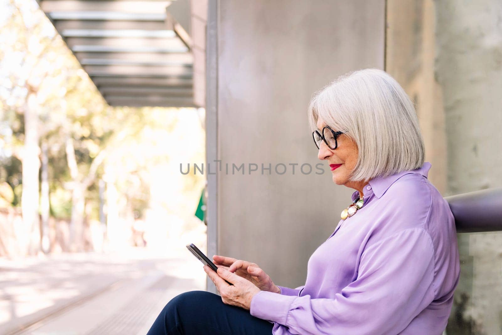 senior woman waiting sitting at bus stop using app on mobile phone, concept of technology and elderly people leisure, copy space for text