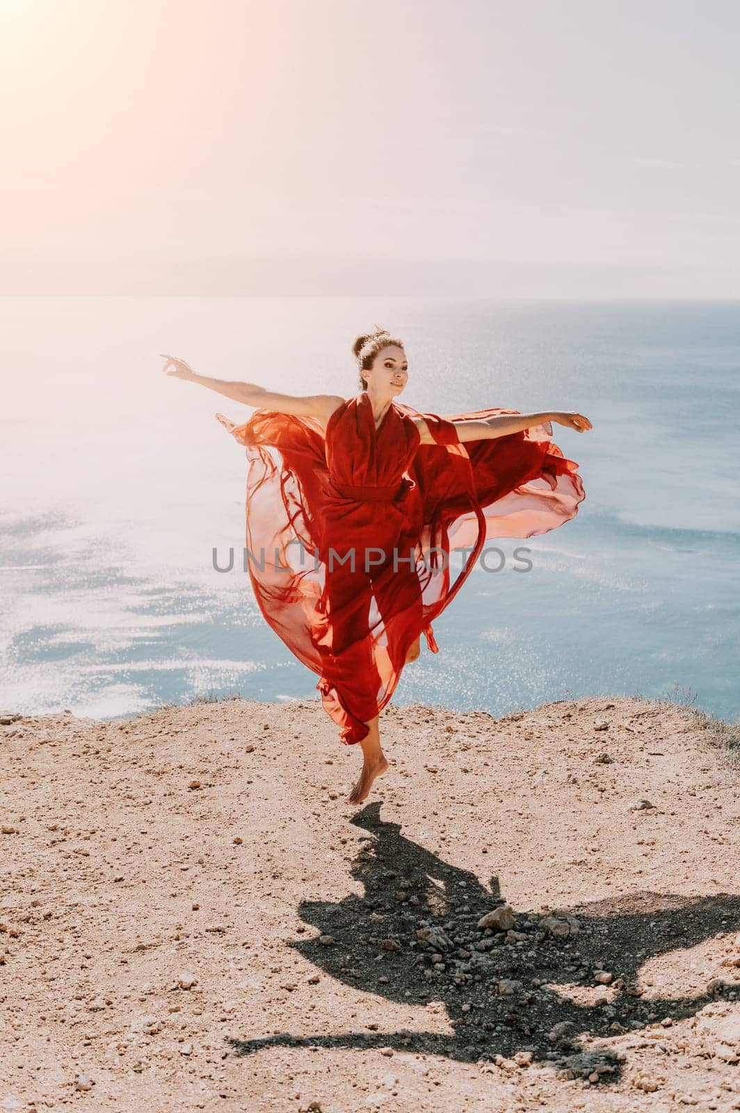 Side view a Young beautiful sensual woman in a red long dress posing on a rock high above the sea during sunrise. Girl on the nature on blue sky background. Fashion photo.