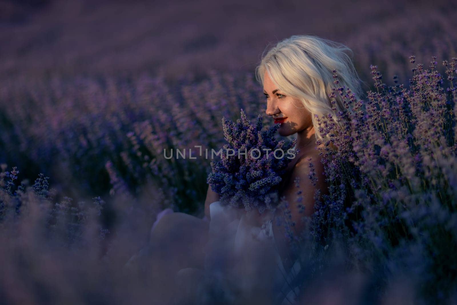 Blonde woman poses in lavender field at sunset. Happy woman in white dress holds lavender bouquet. Aromatherapy concept, lavender oil, photo session in lavender.