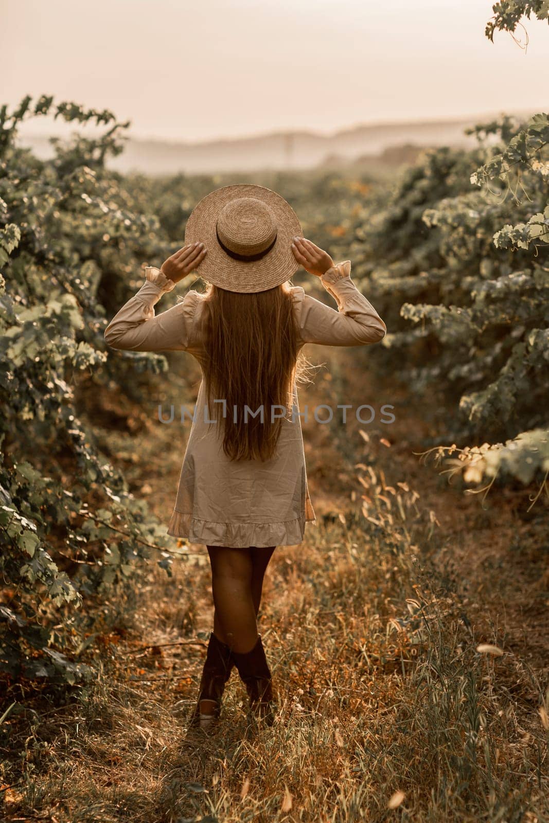 Woman with straw hat stands in front of vineyard. She is wearing a light dress and posing for a photo. Travel concept to different countries.