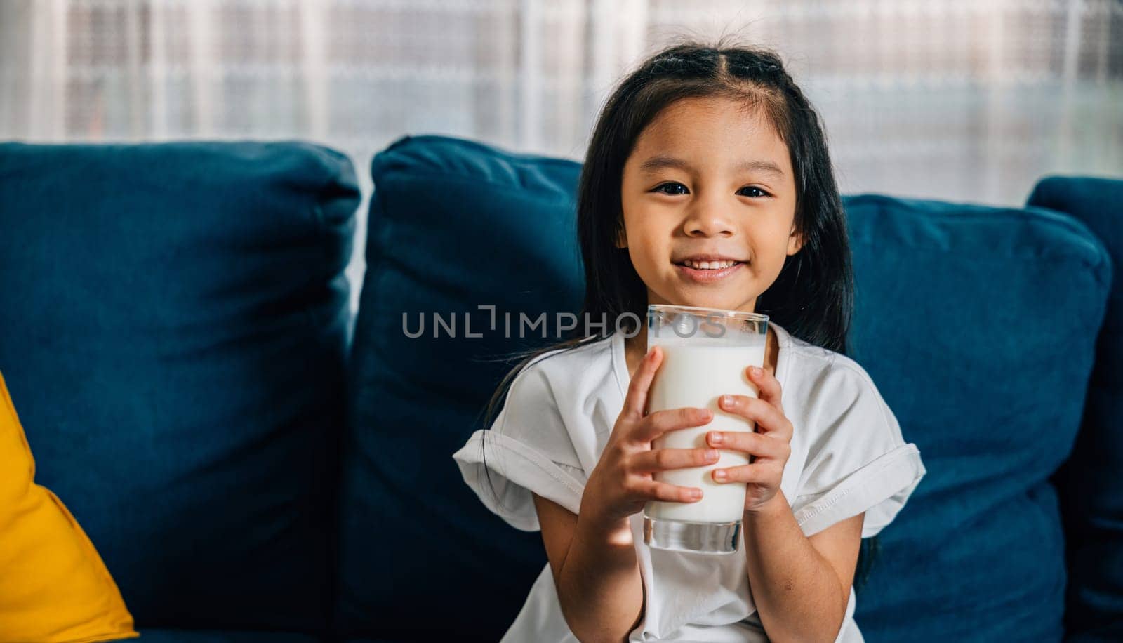 Asian child enjoys a glass of milk on the sofa their happiness and innocence shining through. This photo beautifully represents the significance of daily health care and proper nutrition for children.