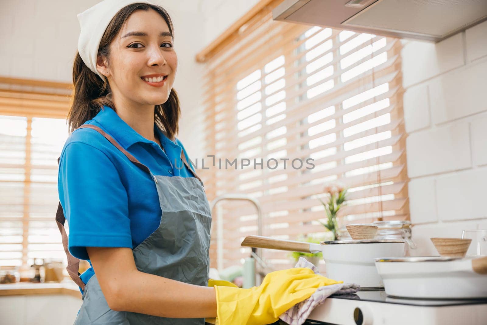 Asian woman in protective gloves wipes dust off gas stove in kitchen. Emphasizing hygiene hand cleaning routine. Cleaner at work safety glove house care. Maid Cleaning housekeeping concept. by Sorapop