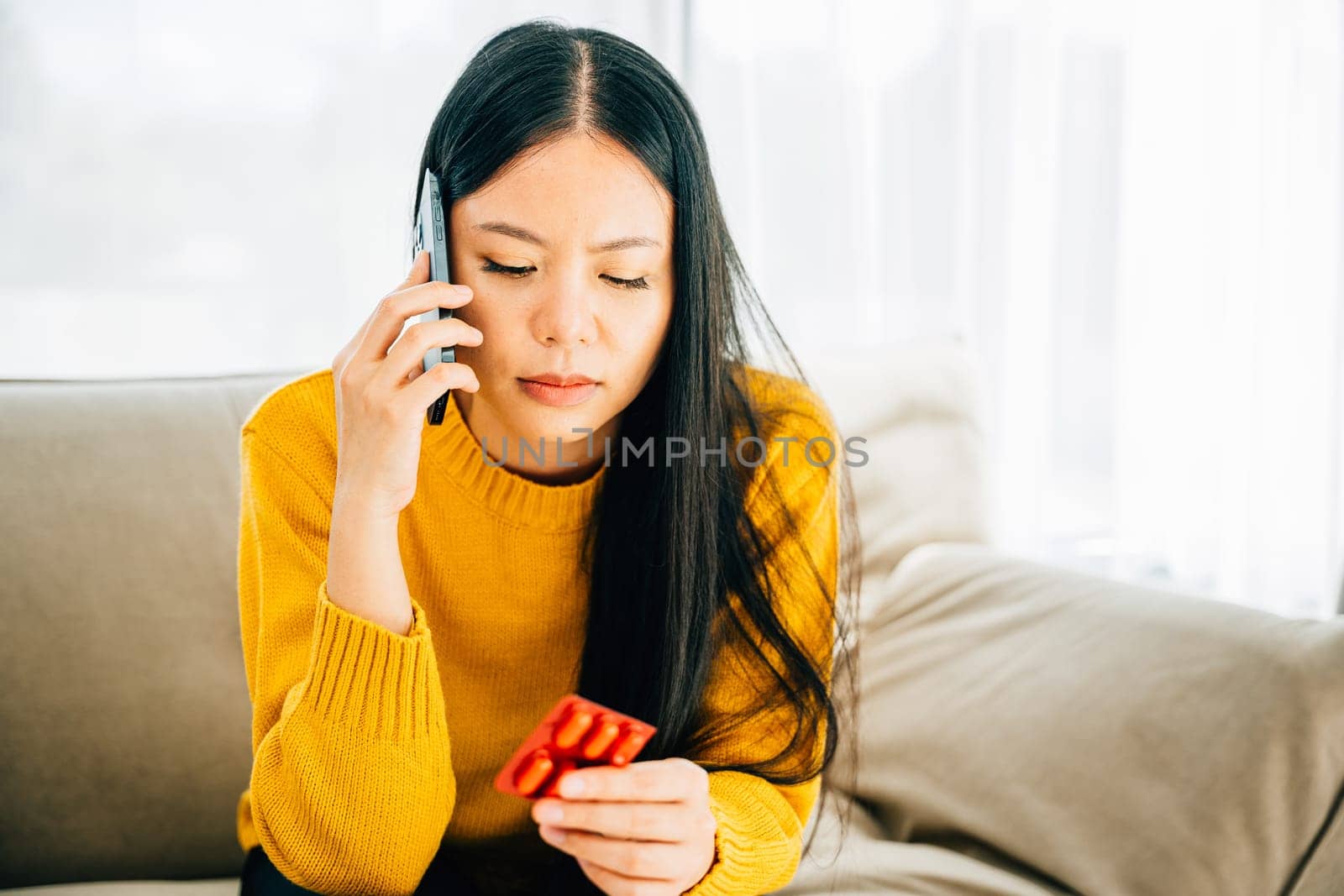 Depicting home consultation, Sick Asian woman on couch taking medicine consulting doctor by phone. Reflecting healthcare quarantine and social distancing due to coronavirus.