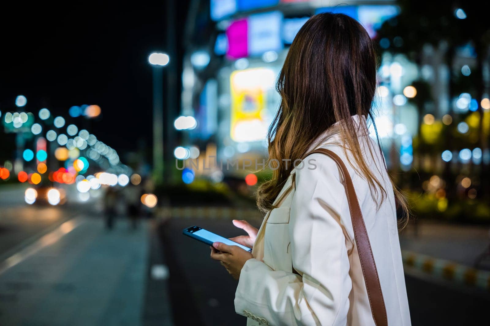 A young woman standing on the side of a city road at night, holding her cellphone and texting, with the glow of city lights in the background.